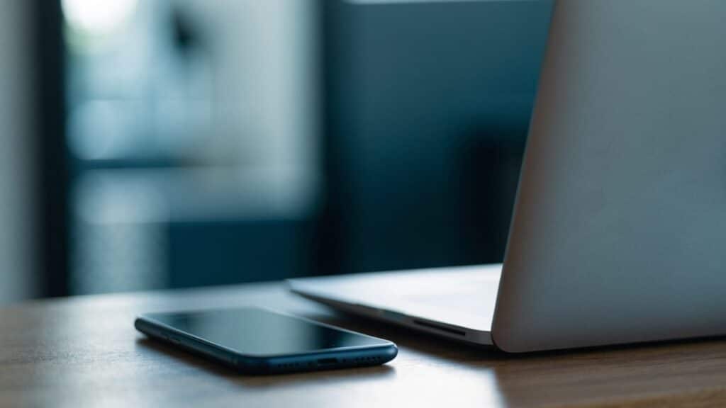 A smartphone lying beside a matte grey laptop on a wooden desk
