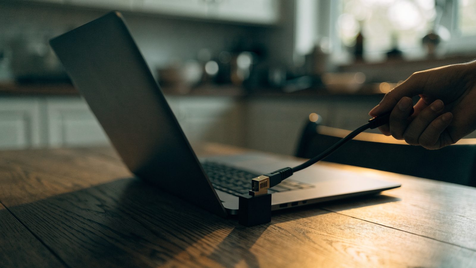 An ethernet cable being plugged into a USB-C adapter connected to a graphite black laptop on a kitchen table
