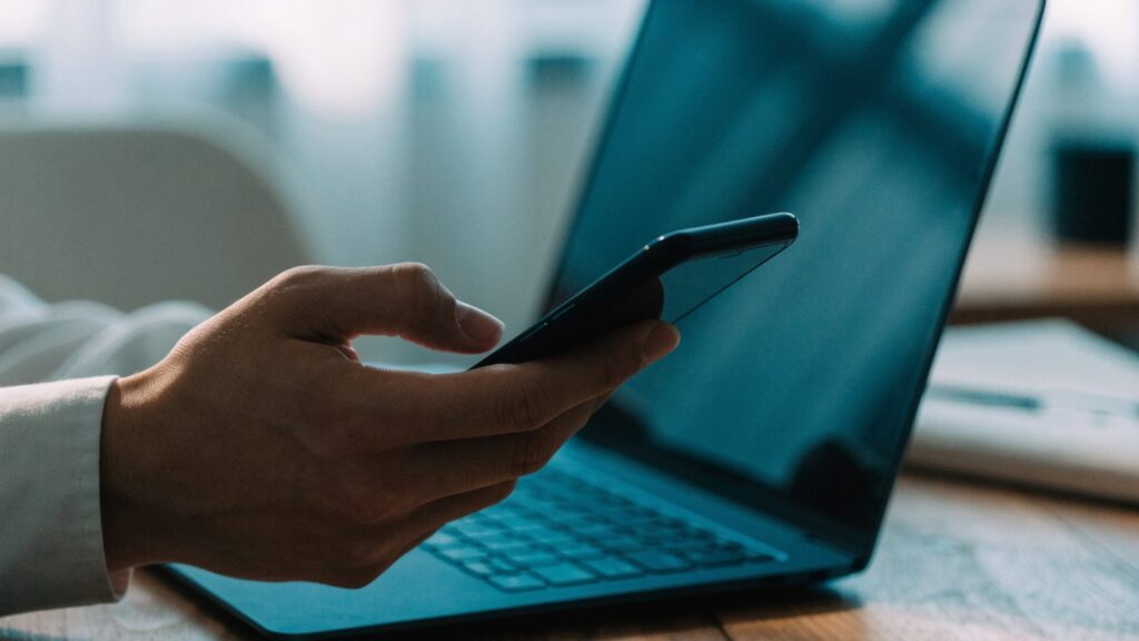 A hand holding a smartphone in front of a dark blue laptop on a wooden desk