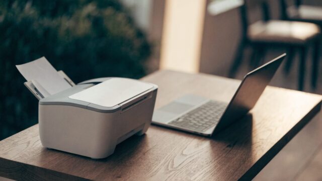 A compact home inkjet printer beside a matte grey laptop on a wooden desk