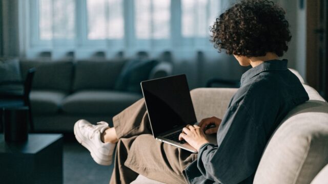 A person seen from behind using a silver laptop on a sofa