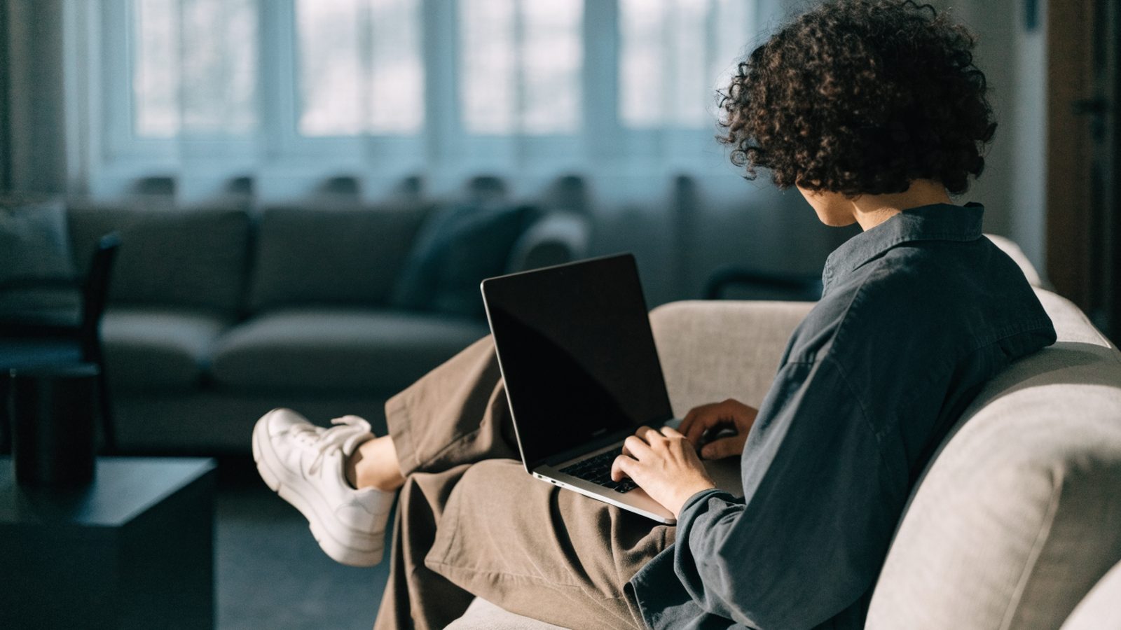 A person seen from behind using a silver laptop on a sofa