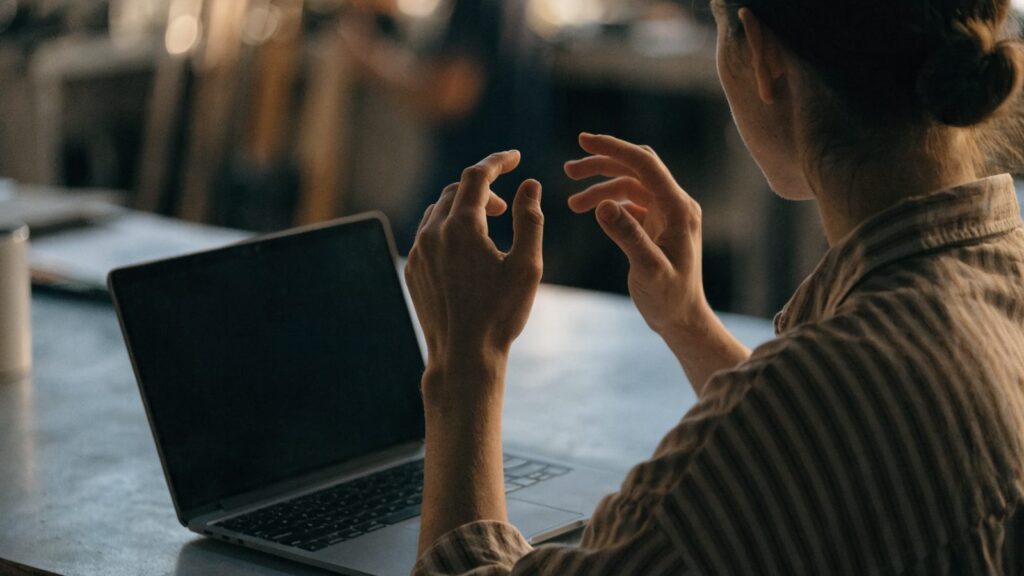 A person seen from behind with hands raised at a matte grey laptop with a dark blank screen on a workbench