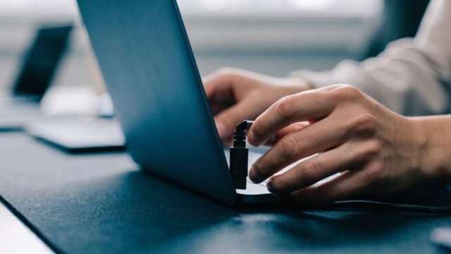 Hands unplugging a charging cable from a slim dark blue laptop on a dark desk mat