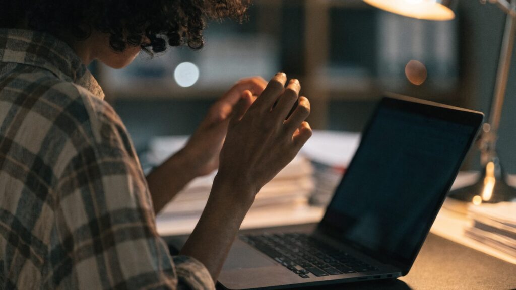 A person seen from behind with hands raised at a graphite black laptop with a dark blank screen on a dark desk
