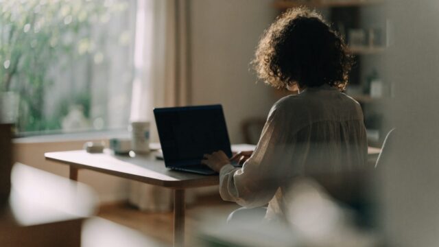 A person seen from behind working on a dark blue laptop in a bright home office