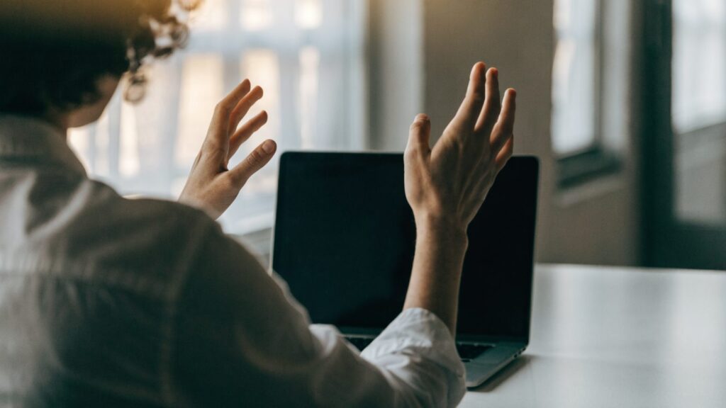 A person seen from behind with hands raised at a matte grey laptop with a dark blank screen on a white desk