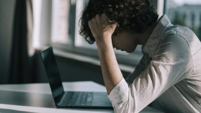 A person resting their forehead on one hand in front of a graphite black laptop on a white desk