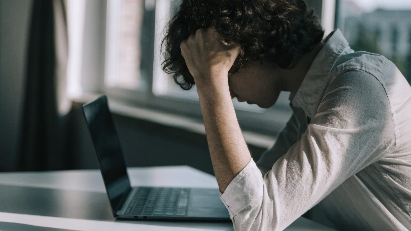A person resting their forehead on one hand in front of a graphite black laptop on a white desk
