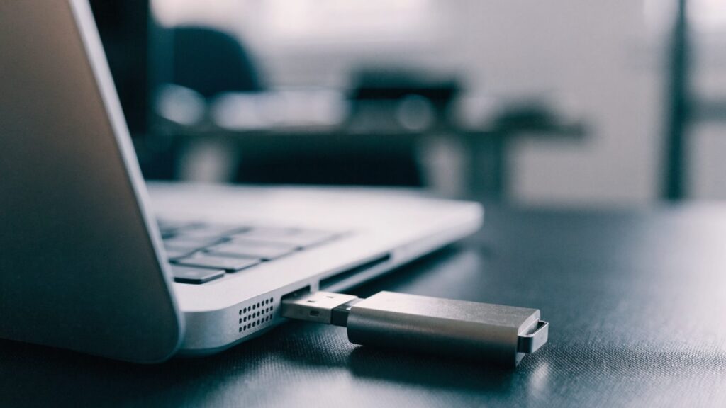 A USB flash drive plugged into the side port of a silver laptop on a dark desk mat