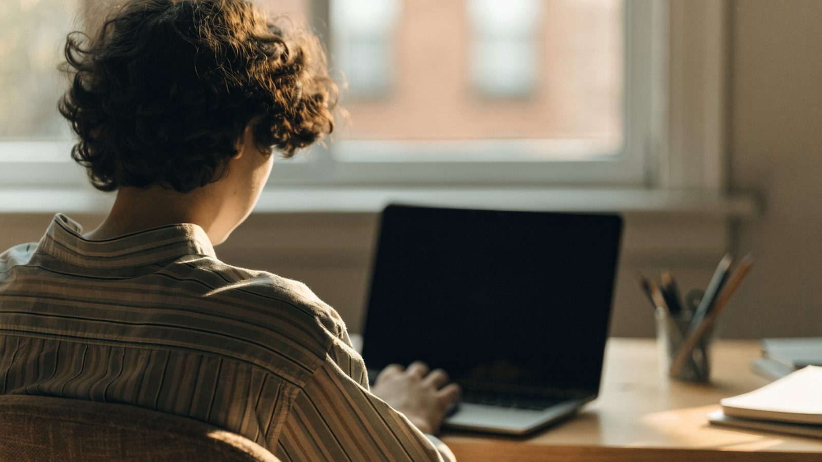 A teenager seen from behind doing homework on a graphite black laptop at a desk