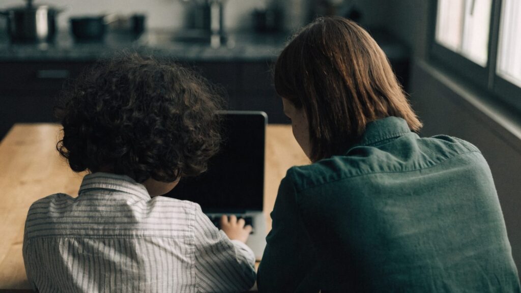 A parent and child seen from behind using a silver laptop together at a kitchen table