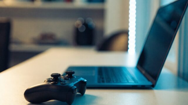 A game controller resting on a desk beside a dark blue laptop in a room with soft LED glow