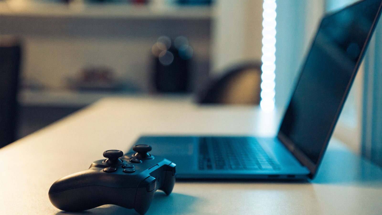 A game controller resting on a desk beside a dark blue laptop in a room with soft LED glow