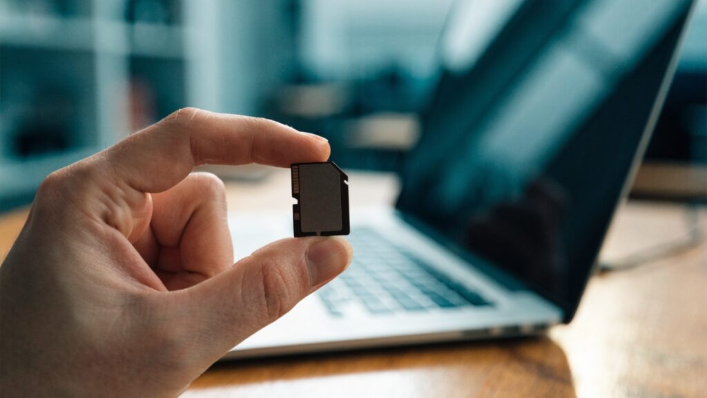 An SD card held between two fingers in front of a silver laptop on a wooden desk