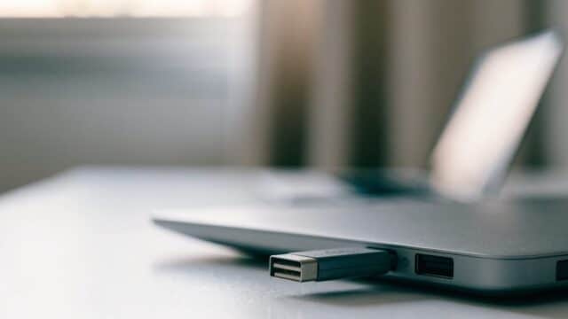 A USB flash drive plugged into the side port of a matte grey laptop on a white desk