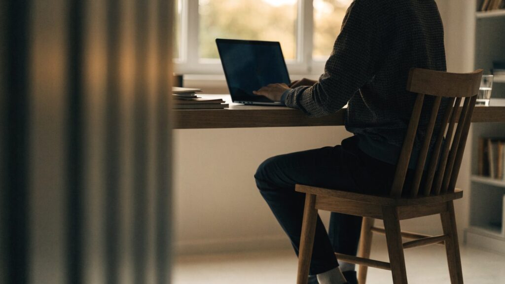A person seen from behind working on a dark blue laptop in a bright home office