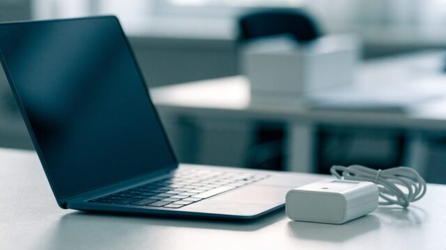 A dark blue laptop on an office desk next to a compact white power adapter with its cable neatly coiled