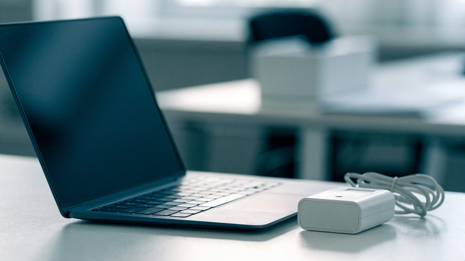 A dark blue laptop on an office desk next to a compact white power adapter with its cable neatly coiled