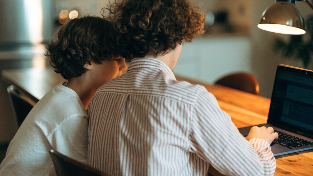 A parent and child seen from behind using a dark blue laptop together at a kitchen table