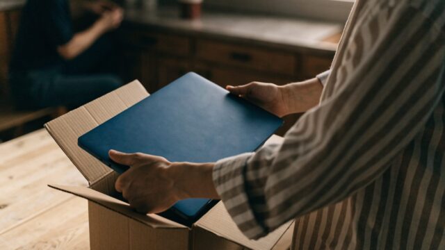 Hands lifting a brand-new dark blue laptop out of its plain cardboard box on a kitchen table