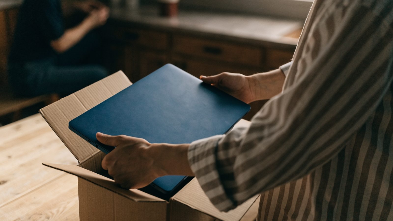 Hands lifting a brand-new dark blue laptop out of its plain cardboard box on a kitchen table