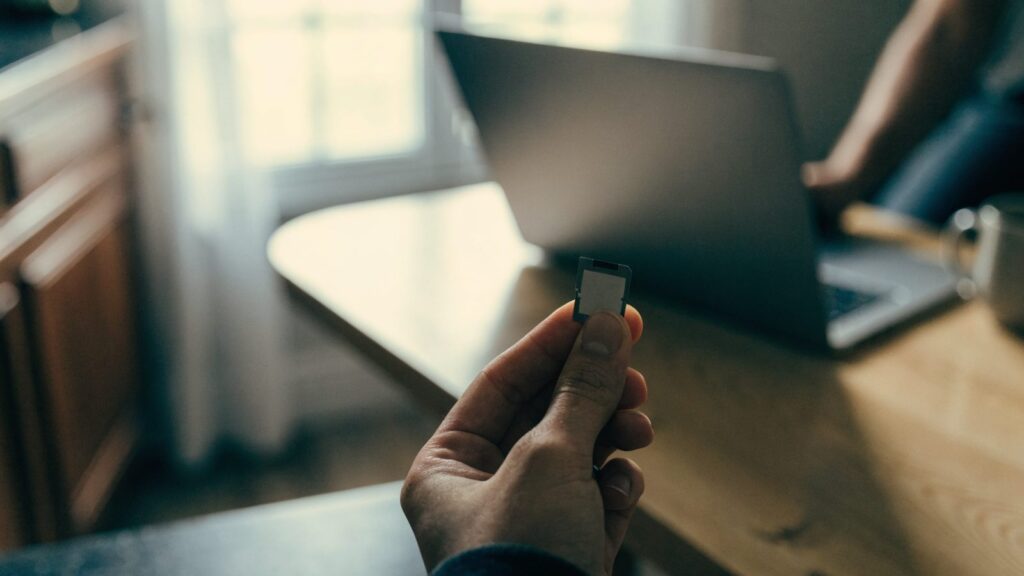 An SD card held between two fingers in front of a matte grey laptop on a kitchen table