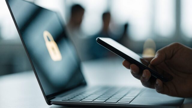 A hand holding a smartphone beside a graphite black laptop