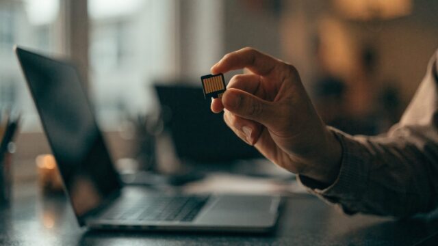 An SD card held between two fingers in front of a matte grey laptop on an office desk