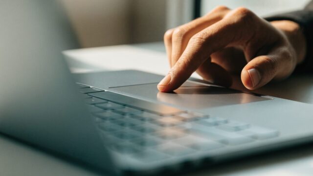 A finger gliding on the touchpad of a matte grey laptop on a white desk