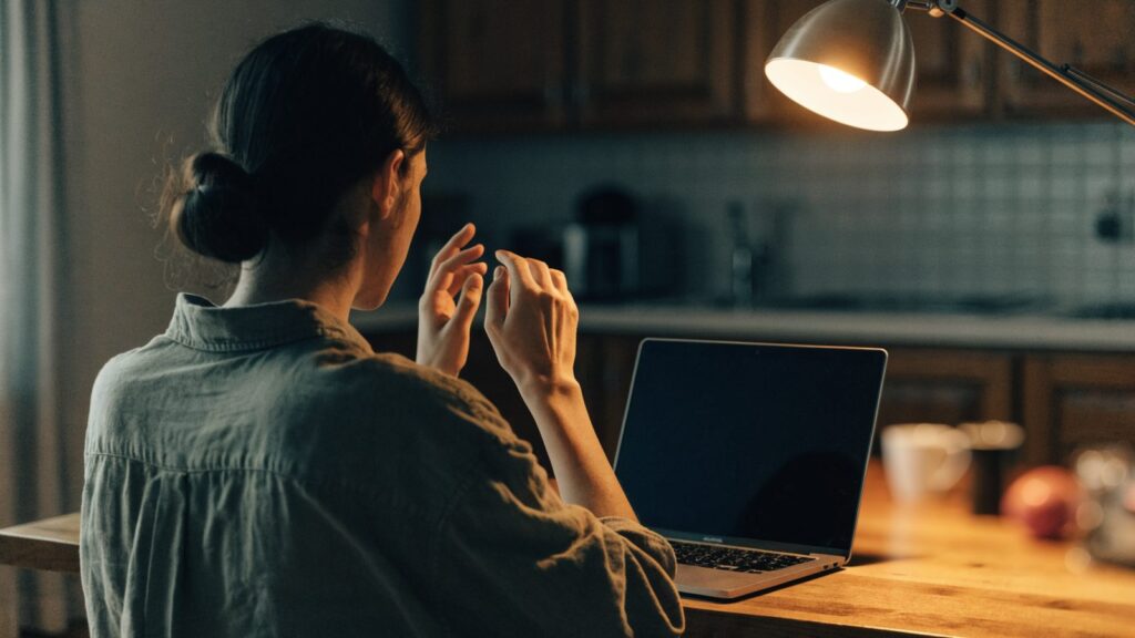 A person seen from behind with hands raised at a matte grey laptop with a dark blank screen on a kitchen table