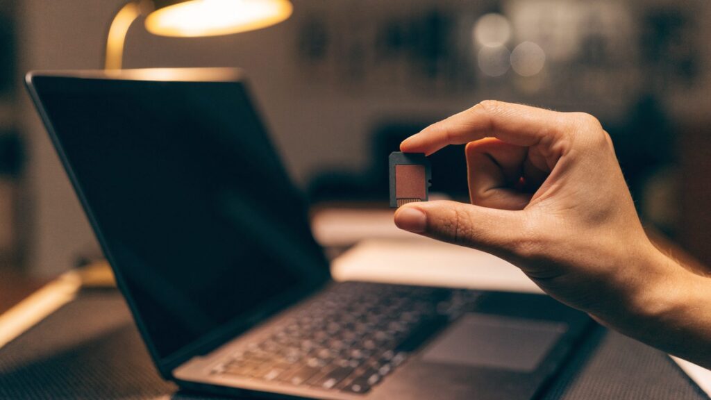 An SD card held between two fingers in front of a graphite black laptop on a dark desk mat