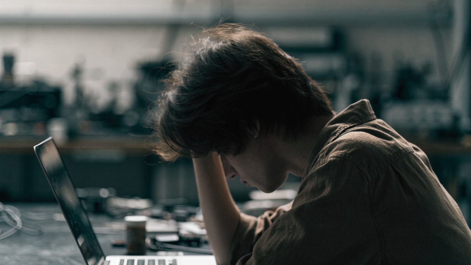 A person resting their forehead on one hand in front of a silver laptop on a workbench