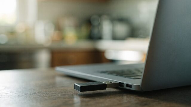 A USB flash drive plugged into the side port of a matte grey laptop on a kitchen table
