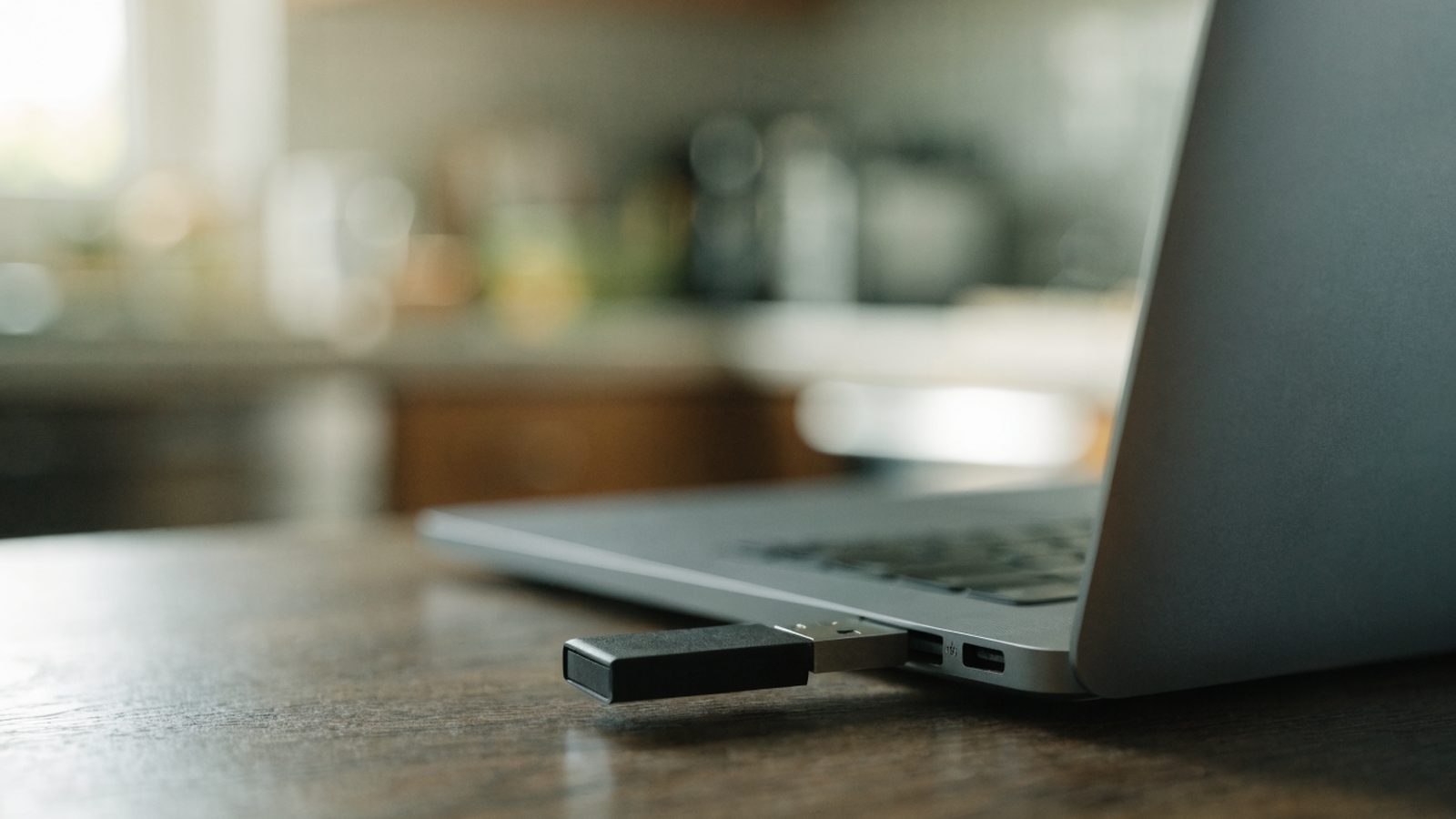 A USB flash drive plugged into the side port of a matte grey laptop on a kitchen table