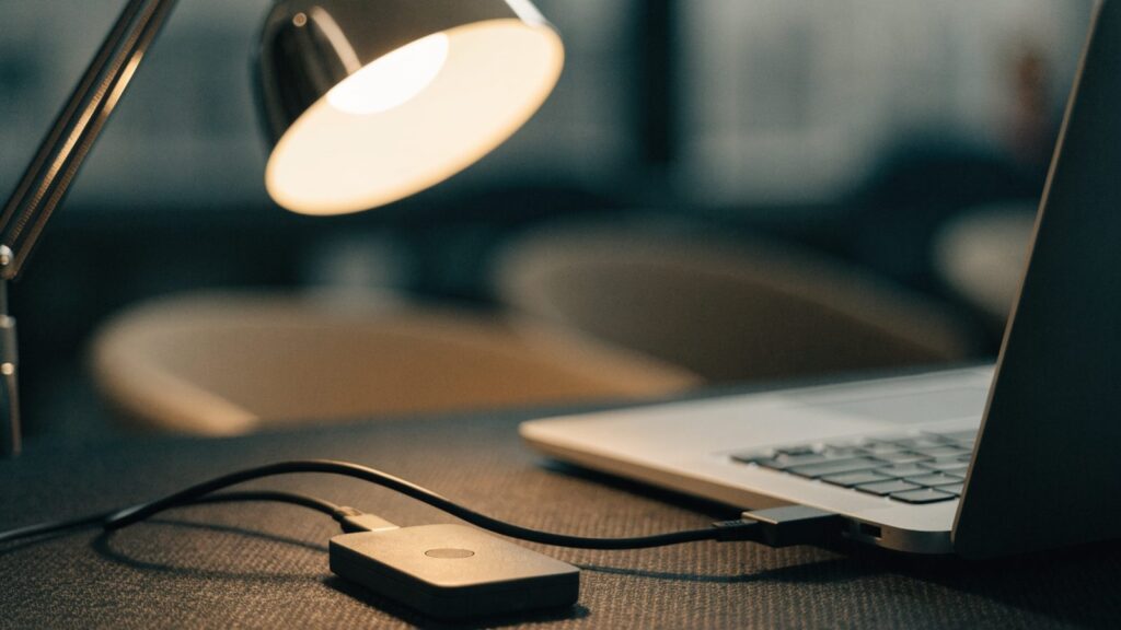 A small external hard drive connected by cable to a silver laptop on a dark desk mat