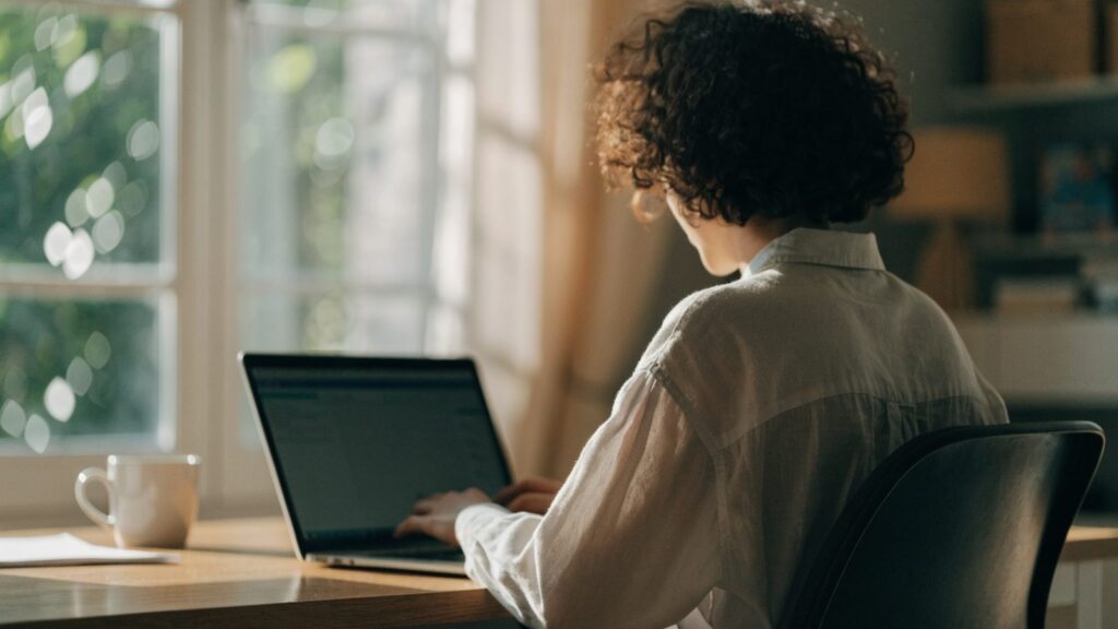 A person seen from behind working on a silver laptop in a bright home office
