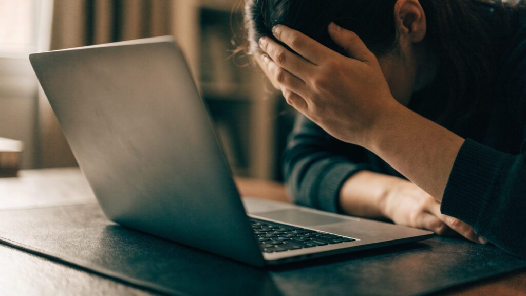 A person resting their forehead on one hand in front of a matte grey laptop on a dark desk mat