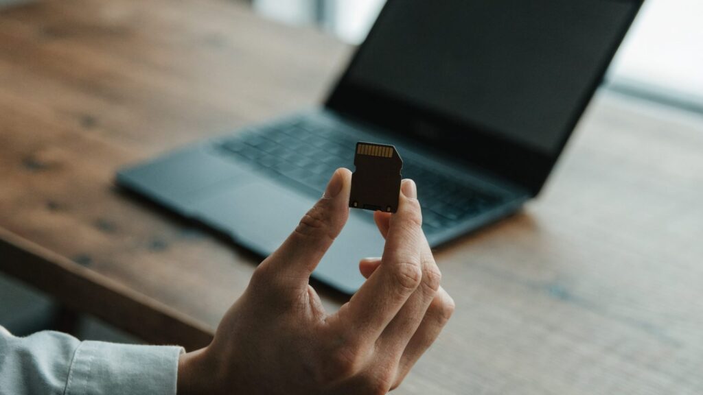 An SD card held between two fingers in front of a graphite black laptop on a wooden desk