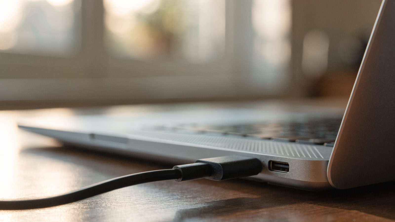 Close-up of a USB-C charger cable plugged into the side port of a silver laptop on a wooden desk
