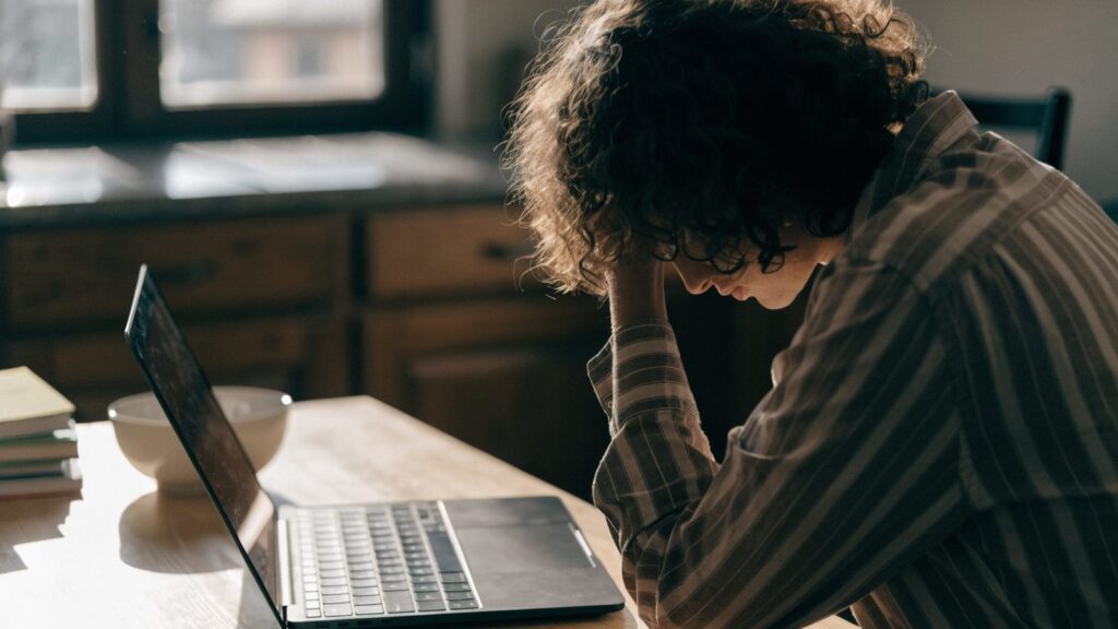 A person resting their forehead on one hand in front of a graphite black laptop on a kitchen table