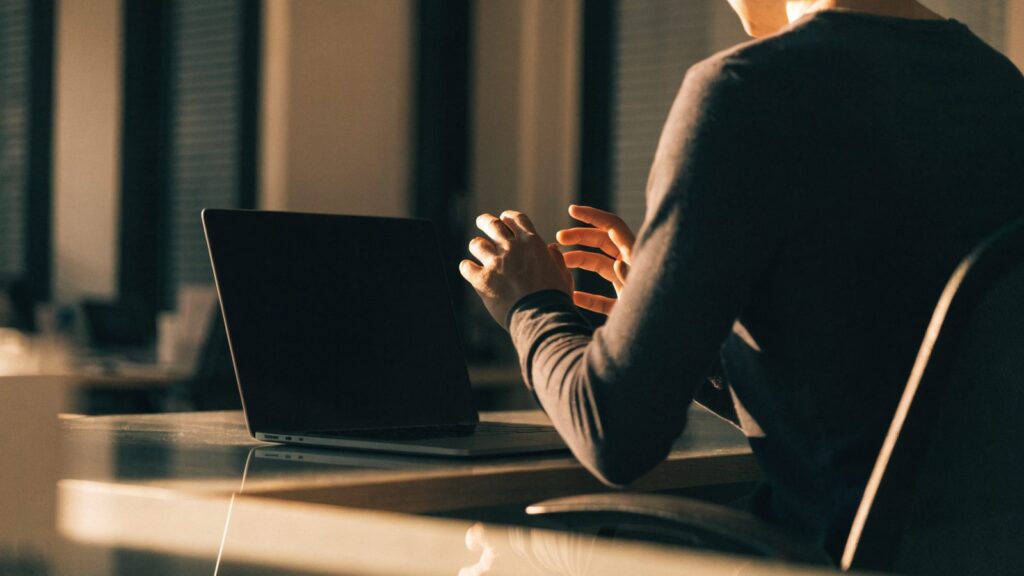 A person seen from behind with hands raised at a graphite black laptop with a dark blank screen on an office d