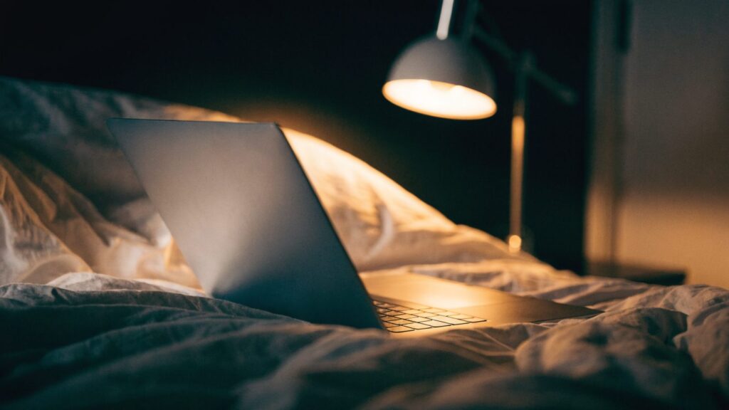 A matte grey laptop glowing softly on a bed in a dark room