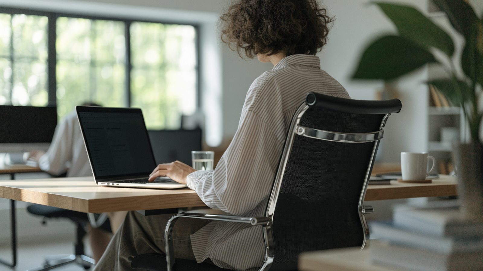 A person seen from behind working on a silver laptop in a bright home office