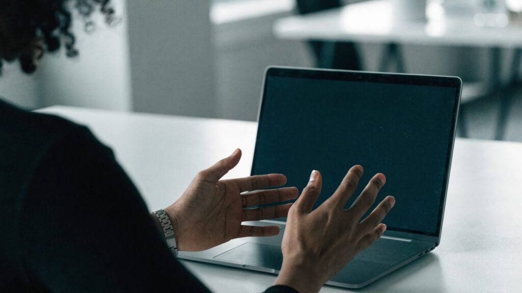 A person seen from behind with hands raised at a matte grey laptop with a dark blank screen on a white desk