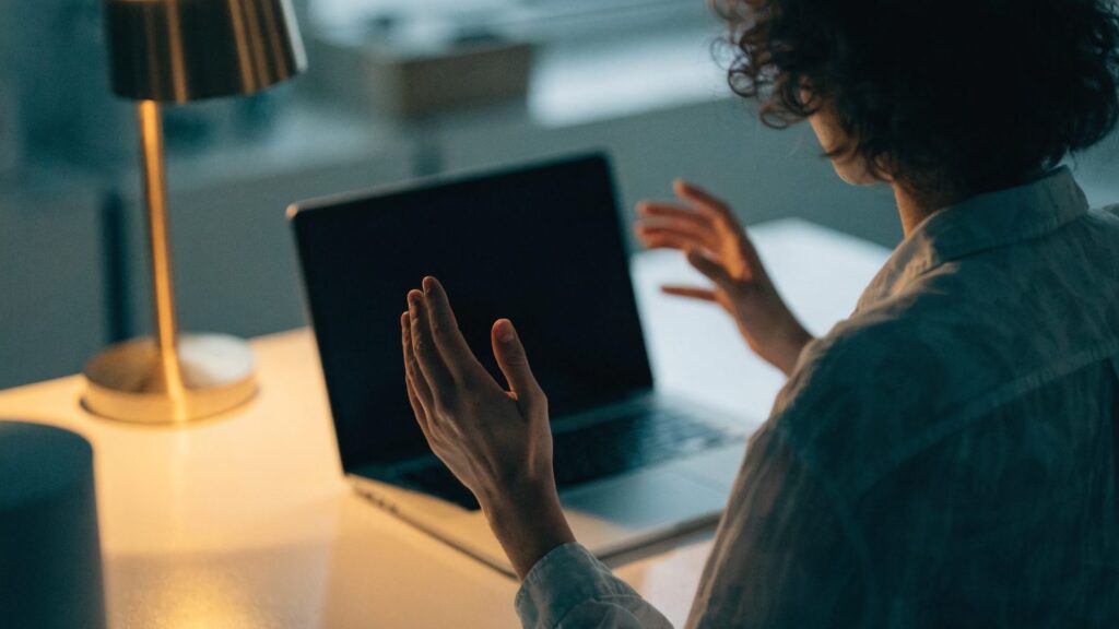 A person seen from behind with hands raised at a silver laptop with a dark blank screen on a white desk