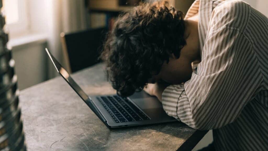 A person resting their forehead on one hand in front of a graphite black laptop on a workbench
