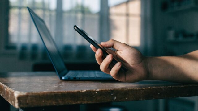A hand holding a smartphone in front of a dark blue laptop on a workbench