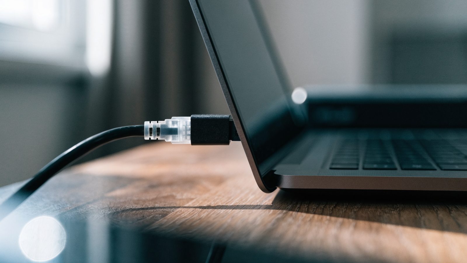 An ethernet cable being plugged into a USB-C adapter connected to a graphite black laptop on a wooden desk
