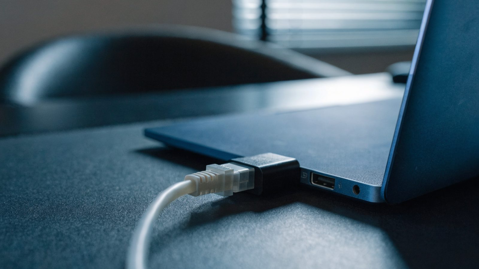 An ethernet cable being plugged into a USB-C adapter connected to a dark blue laptop on a dark desk mat
